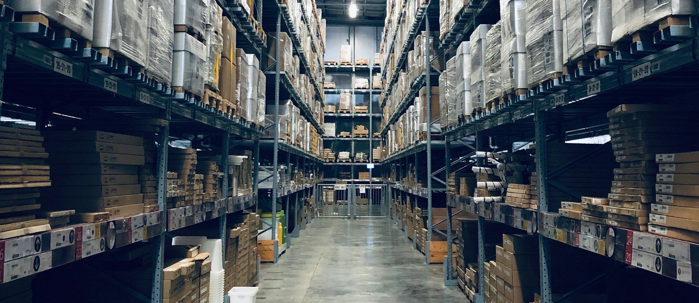 Pasillo central de una bodega con estanterías de altura a ambos lados, cargadas de pallets envueltos y cajas apiladas.