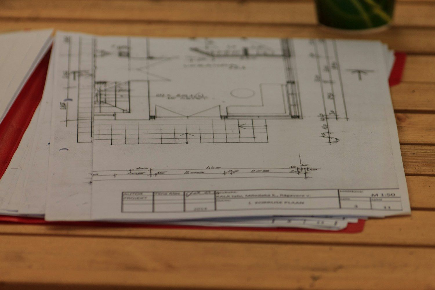Planos de arquitectura acotados a escala 1:50, apilados sobre una carpeta roja en el escritorio de una oficina técnica.