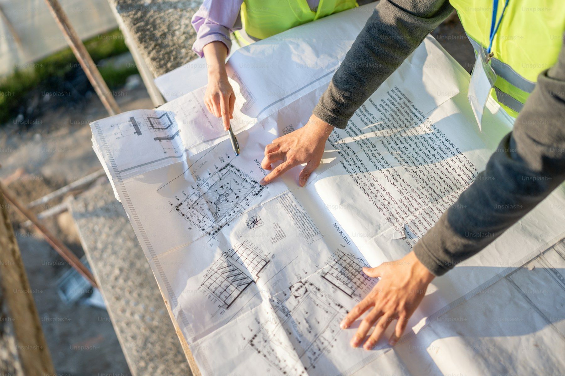 Dos personas con chaleco reflectante revisan un plano de arquitectura desplegado sobre una mesa, en el terreno de una obra.